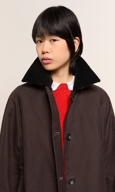 Plümo  Person with short black hair wears the Doherty Coat - Soil with a raised collar and red sweater, looking at the camera.