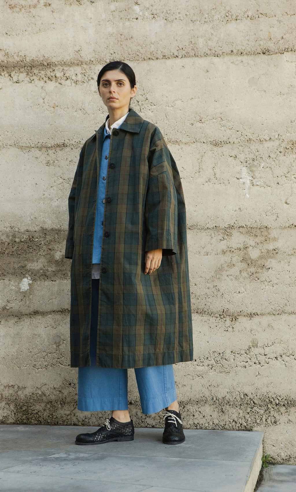 Plümo- Person in a Doherty Coat - Tartan, denim pants, and black shoes stands by a textured concrete wall.