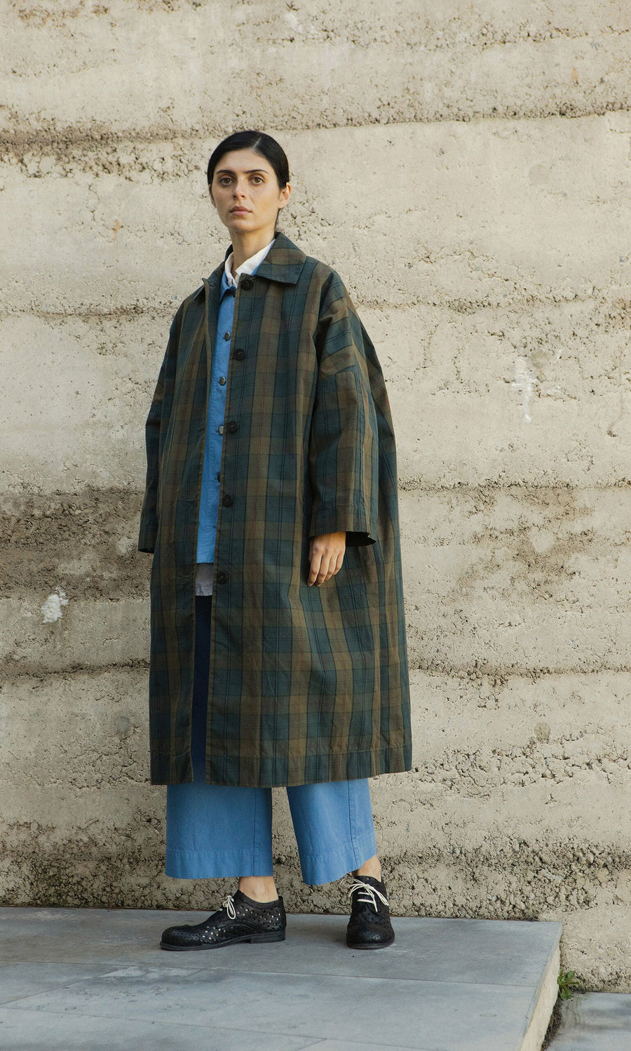 Plümo- Person in a Doherty Coat - Tartan, denim pants, and black shoes stands by a textured concrete wall.