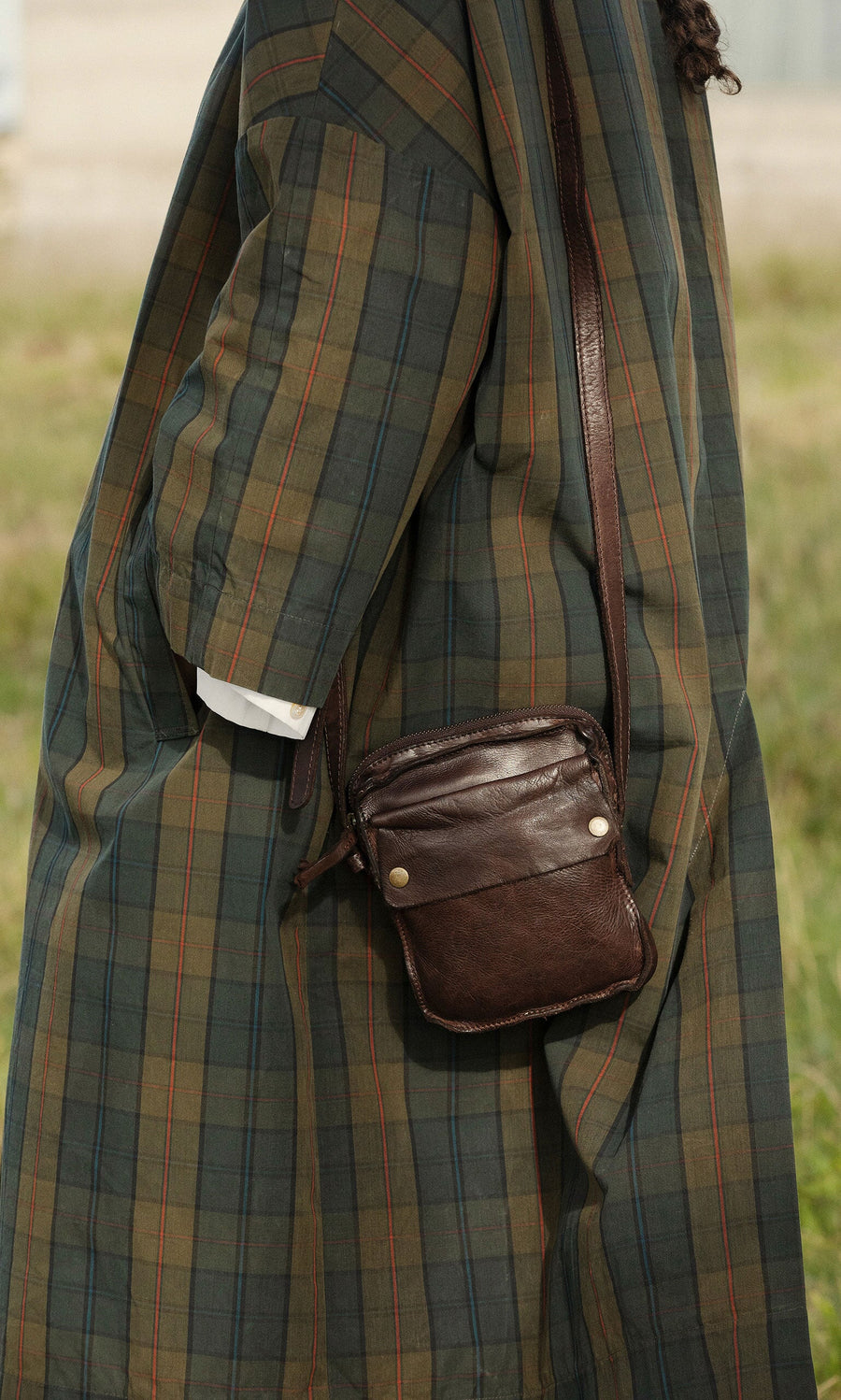 Plümo- Person in a green plaid coat with the Orton Parcel Bag, standing outdoors on grass, embracing British design.