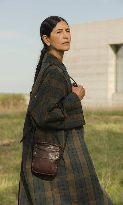 Plümo- A person in the Doherty Coat - Tartan stands outdoors with a brown crossbody bag and a modern building behind them.