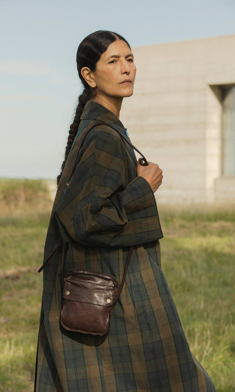 Plümo- A person in the Doherty Coat - Tartan stands outdoors with a brown crossbody bag and a modern building behind them.