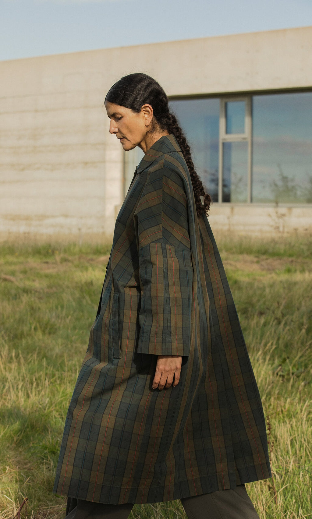 Plümo- A woman with long dark hair in braids wears the Doherty Coat - Tartan, walking outside by a modern building.
