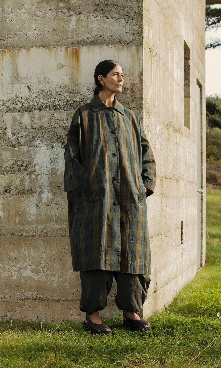 Plümo- Wearing the Doherty Coat - Tartan, a person stands outdoors by a concrete wall, hands in pockets, looking aside.