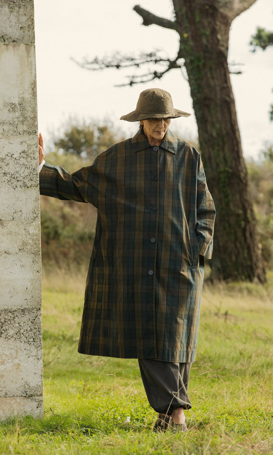 Plümo- An elderly person in a Doherty Coat - Tartan and wide-brimmed hat stands by a concrete pillar in a grassy area.