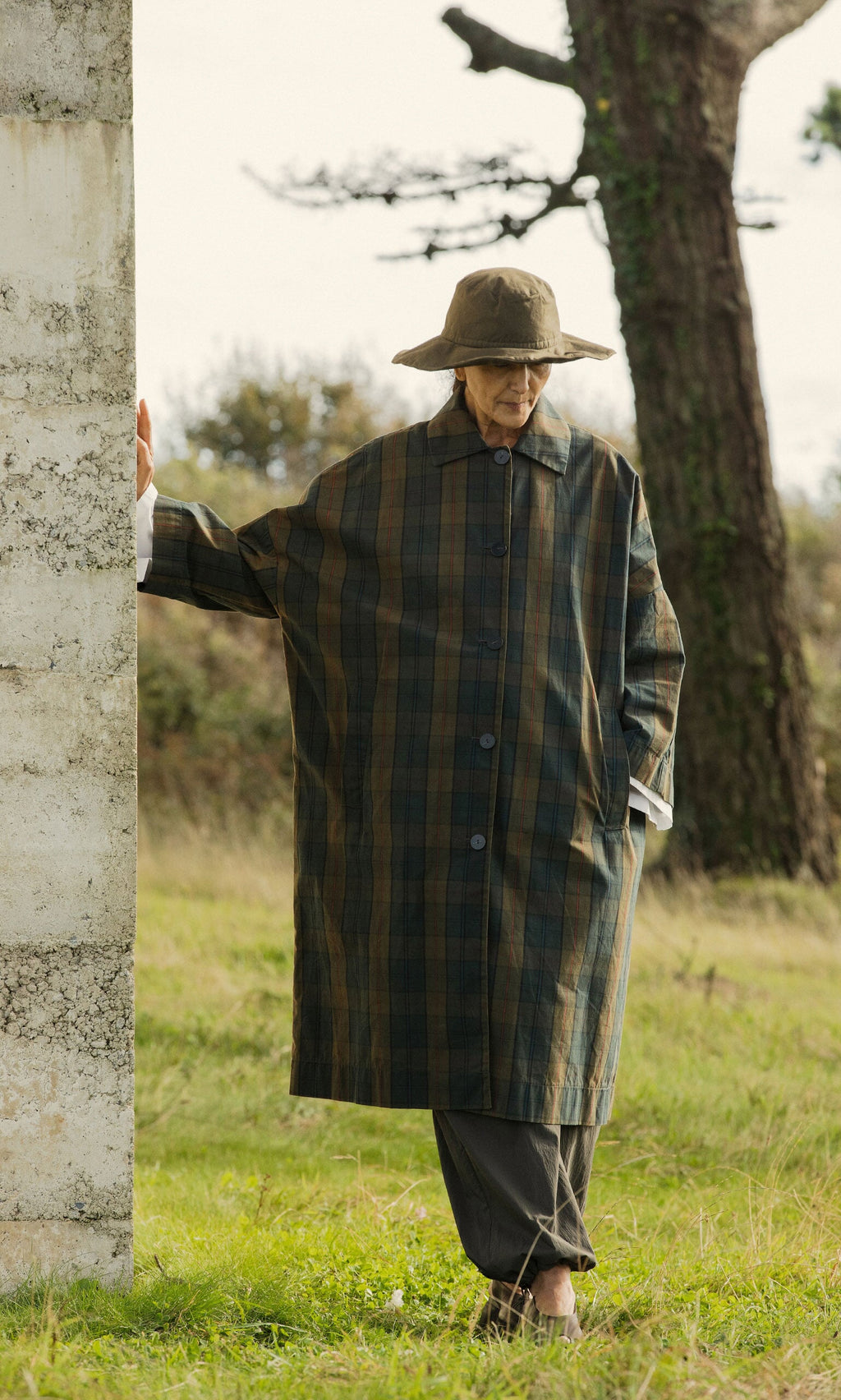 Plümo- An older person in a Gaucho Waxed Hat and plaid coat stands by a stone wall in a grassy outdoor setting.
