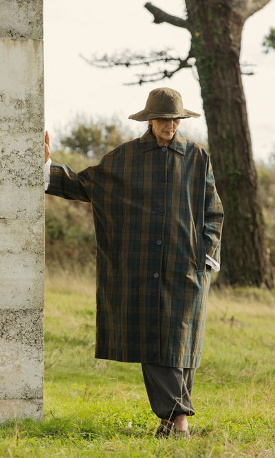 Plümo- An older person in a Gaucho Waxed Hat and plaid coat stands by a stone wall in a grassy outdoor setting.