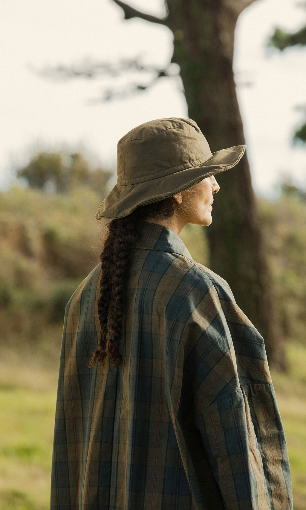 Plümo- A person with braided hair in a waxed cotton jacket and Gaucho Waxed Hat stands outdoors facing sideways.