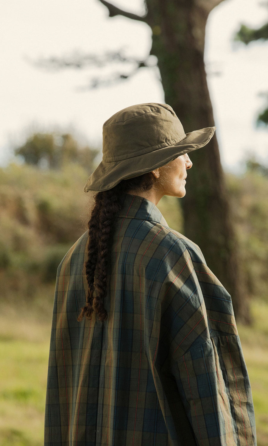 Plümo- A person with braided hair in a waxed cotton jacket and Gaucho Waxed Hat stands outdoors facing sideways.