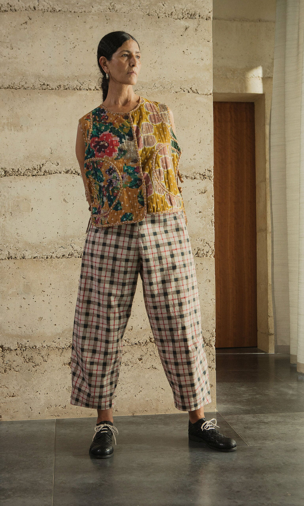 Plümo- A person stands indoors in a floral sleeveless top, Dundee Barrel Pants with a trompe l’oeil check, and black shoes.