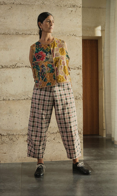 Plümo- A person stands indoors in a floral sleeveless top, Dundee Barrel Pants with a trompe l’oeil check, and black shoes.