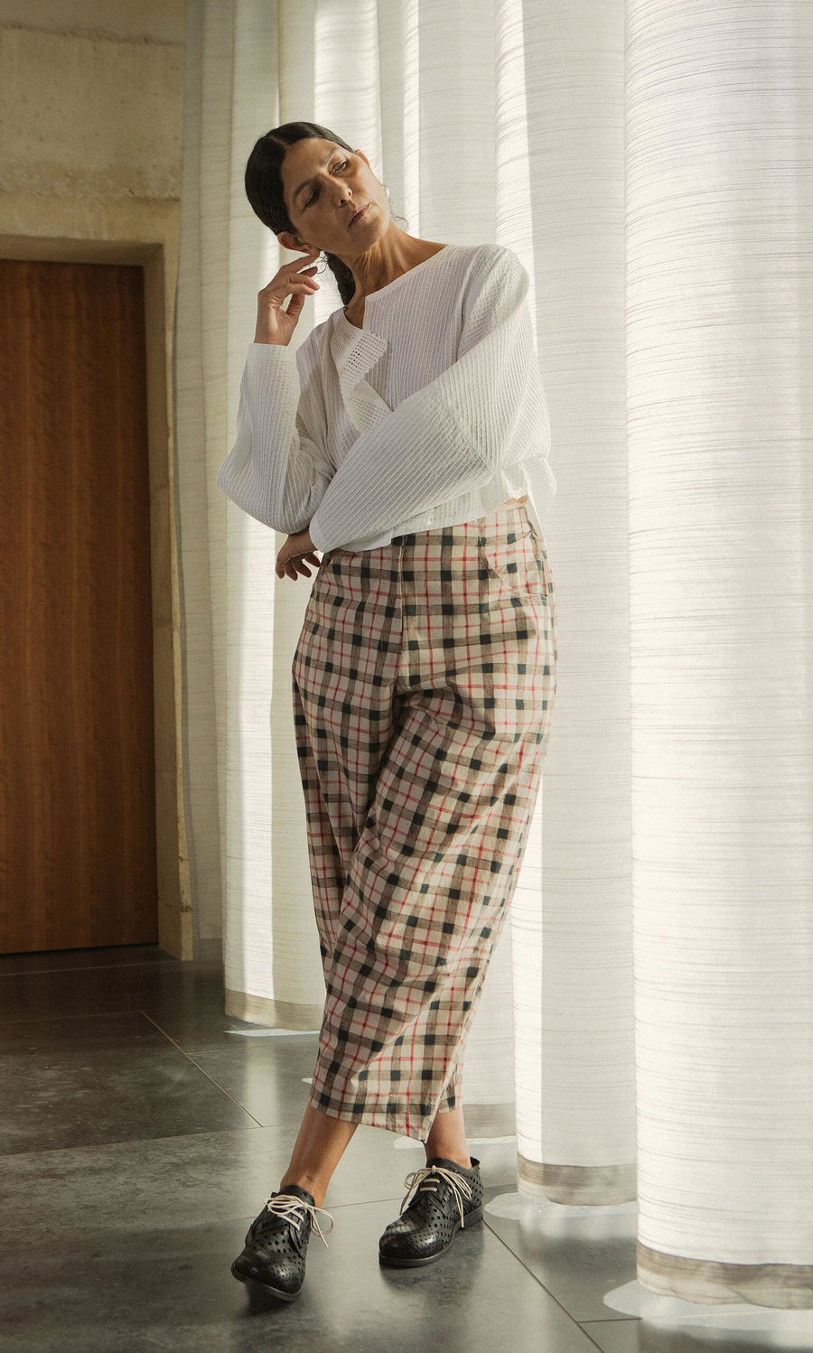 Plümo- Woman in a white blouse and Dundee Barrel Pants stands by sheer curtains, hand near her face.