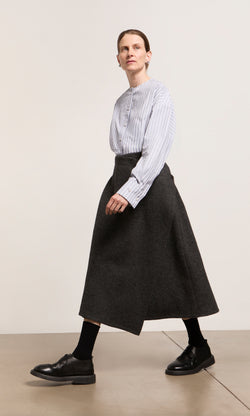 Plümo  Person in a striped blouse, Eardley Skirt, black socks, and shoes walks indoors on a light-colored floor.