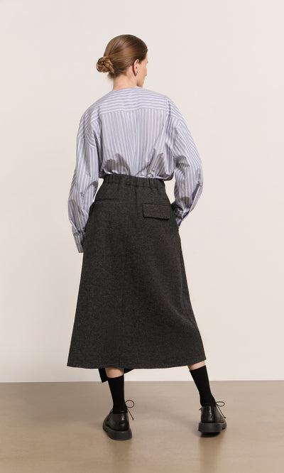 Plümo  Woman faces away in striped shirt, Eardley Skirt, black socks and shoes, set against a plain background.