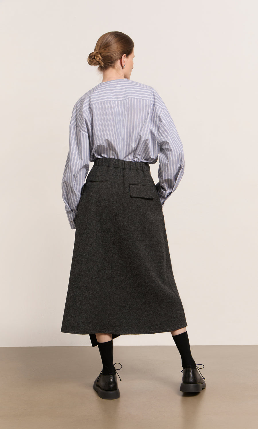 Plümo  Woman faces away in striped shirt, Eardley Skirt, black socks and shoes, set against a plain background.