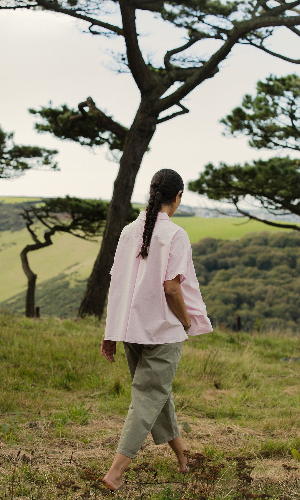 Plümo- A person with a braid walks barefoot in grass, wearing the Early Pink Blouse, with trees and hills behind.