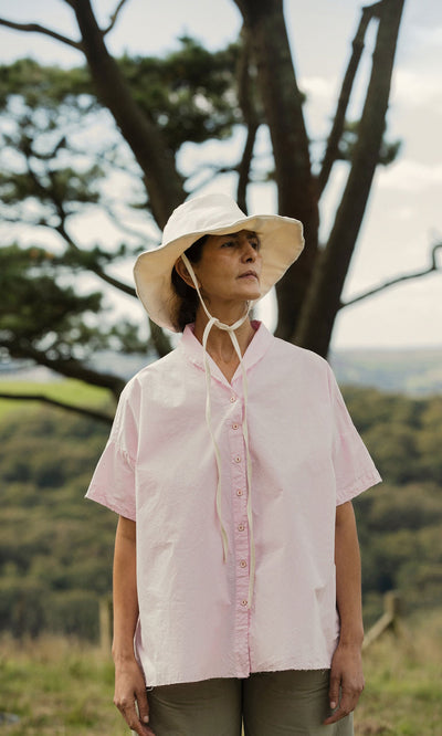 Plümo- A woman in an Early Pink Blouse by Hannoh Wessel stands outdoors with trees and hills in the background.