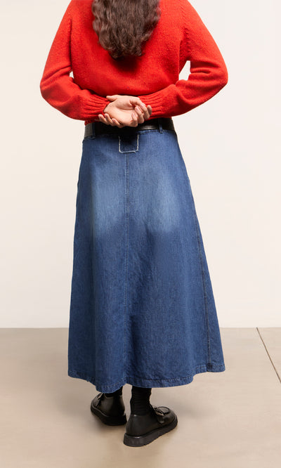 Plümo  Person in a red sweater and Eday Denim Skirt stands with hands behind back, facing away from the camera.