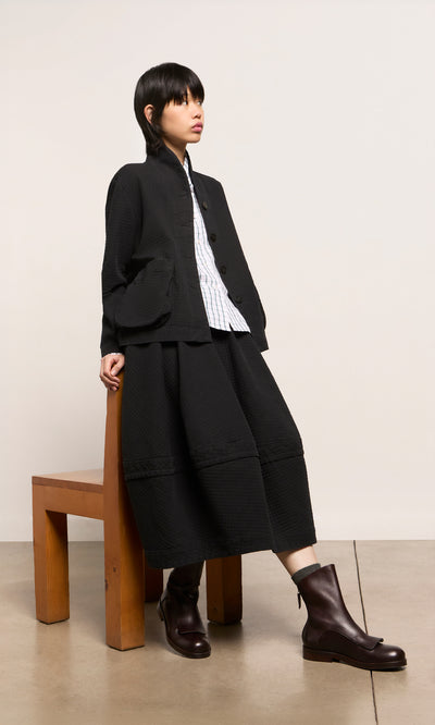 Plümo  Person in an Envoy Jacket - Coal, striped shirt, and brown boots sits on a bench against a plain background.