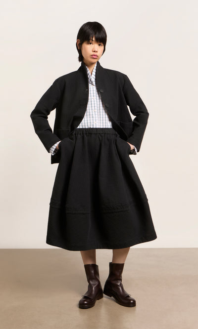 Plümo  Person wears the Envoy Jacket - Coal, a white checkered shirt, skirt, and brown boots; hands in pockets.