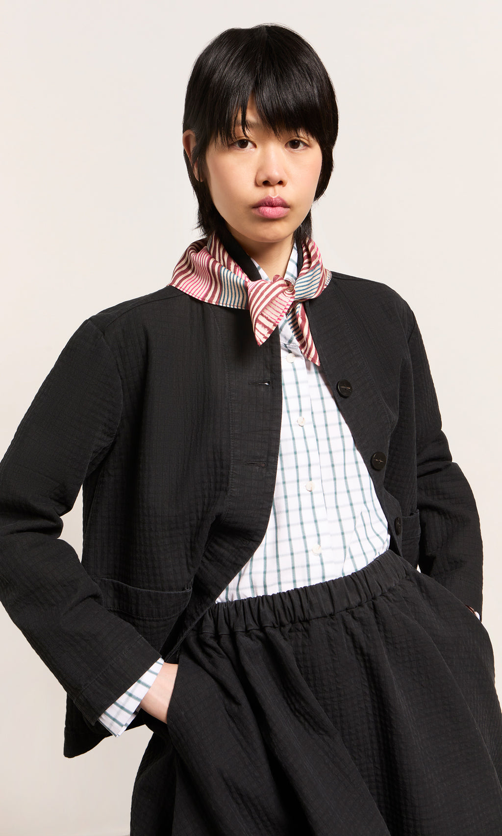 Plümo  Person wears a black jacket, checkered shirt, Match Necktie, and skirt, standing with hand in pocket.
