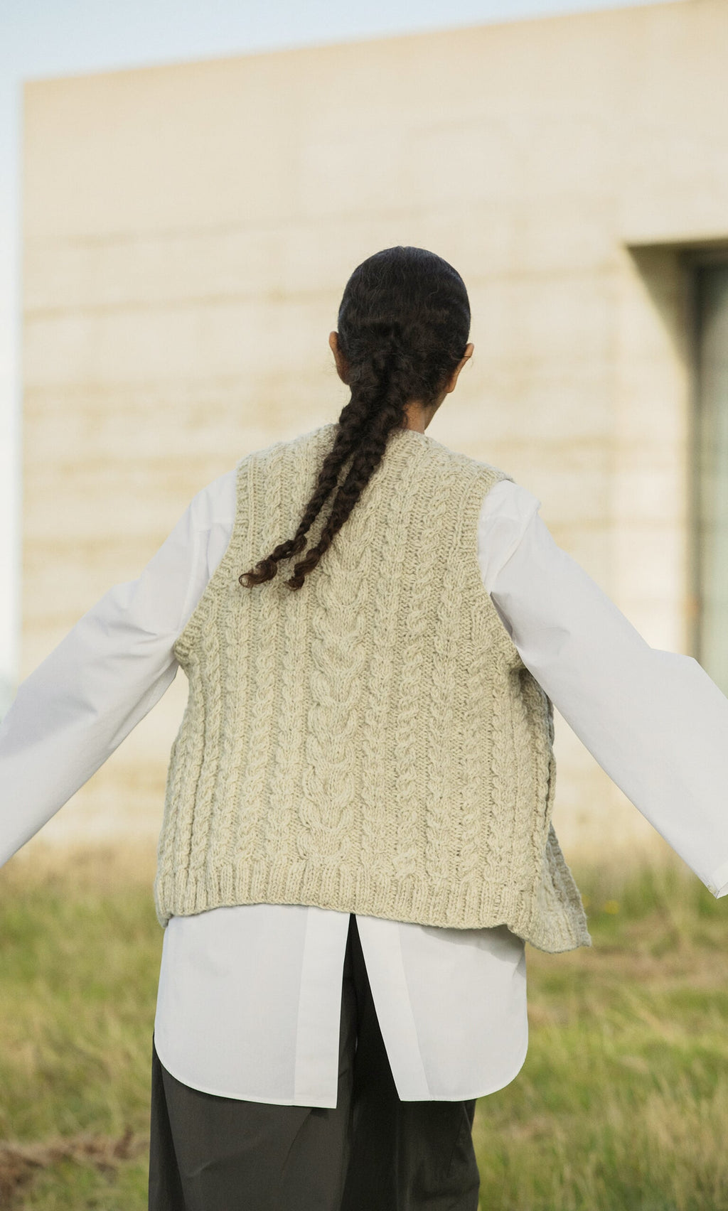 Plümo- Person with braided hair in a Finstock Waistcoat over a white shirt stands outdoors with arms outstretched.
