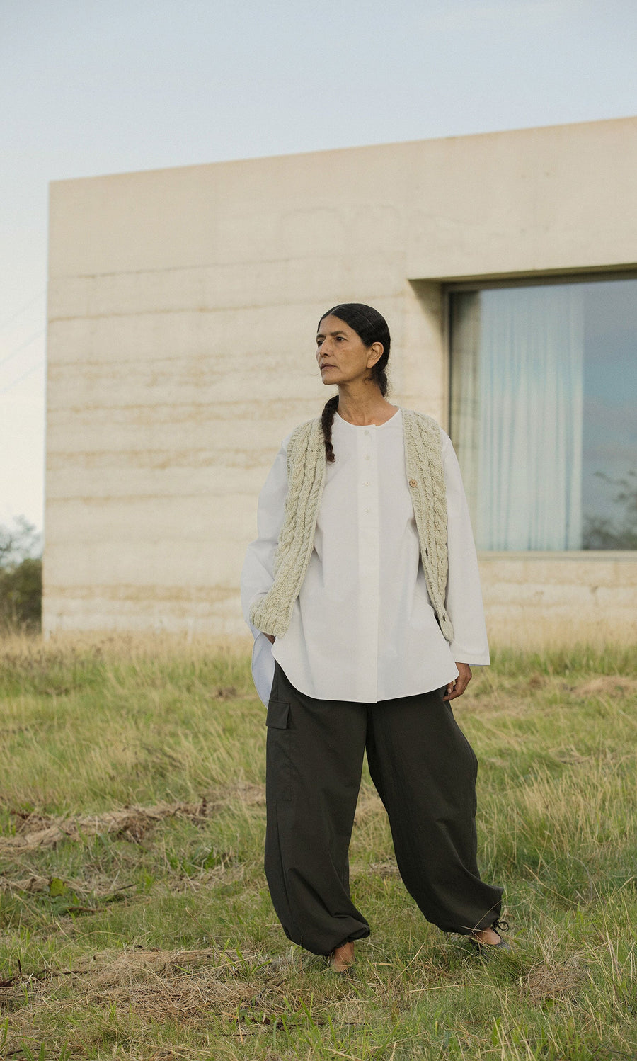 Plümo- Woman in loose white shirt, Finstock Waistcoat, and dark pants stands barefoot on grass by a modern building.