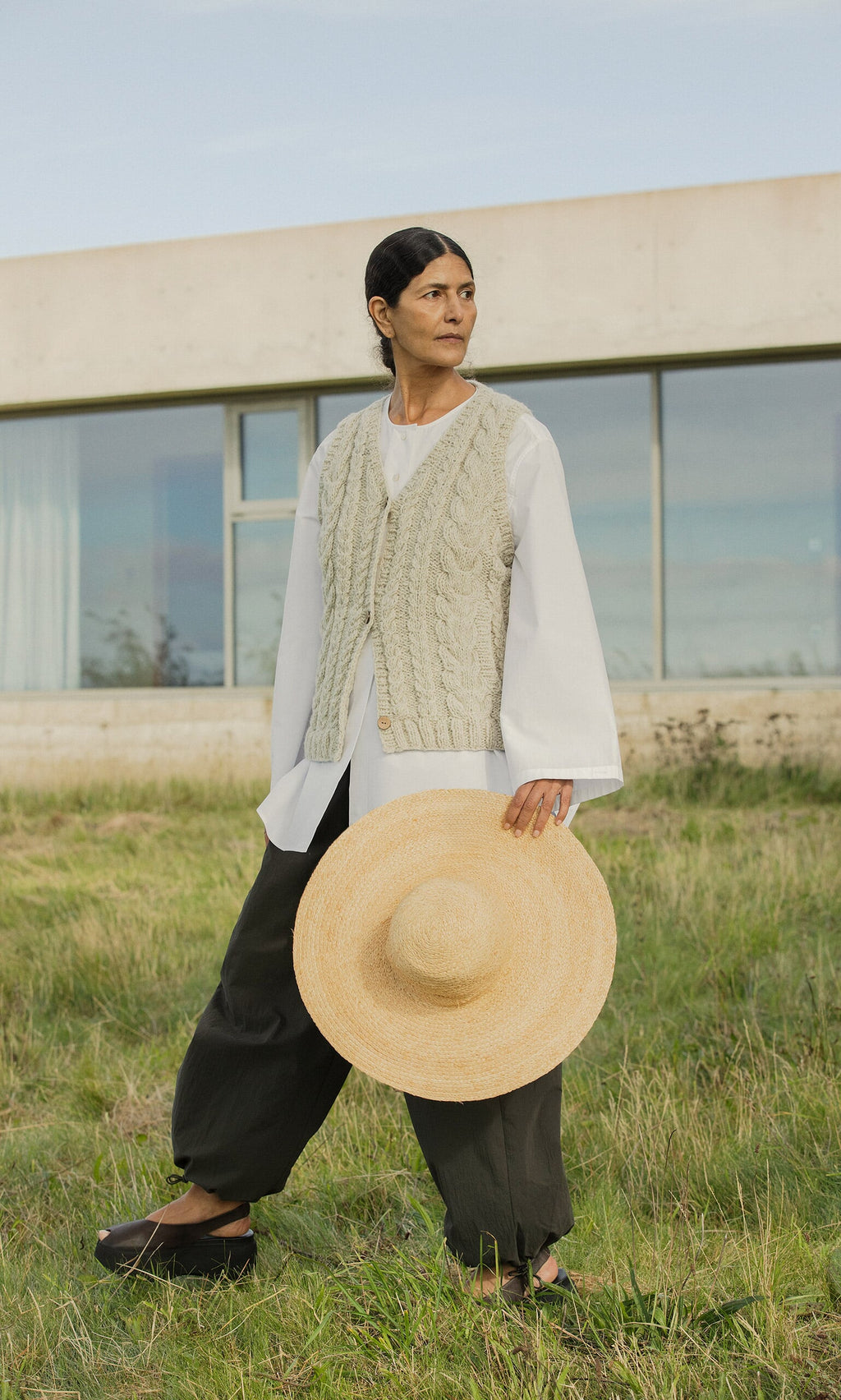 Plümo- On grass, a person in loose pants, white shirt, and Fairtrade Finstock Waistcoat holds a straw hat.