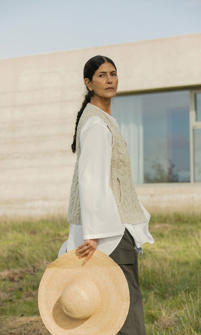 Plümo- A woman in a white shirt and the Finstock Waistcoat holds a straw hat, standing on grass near a modern building.