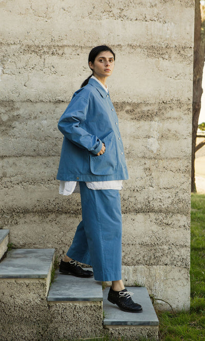 Plümo- A woman in Cromer Pants - French Blue stands on stone steps by a textured wall, looking toward the camera.