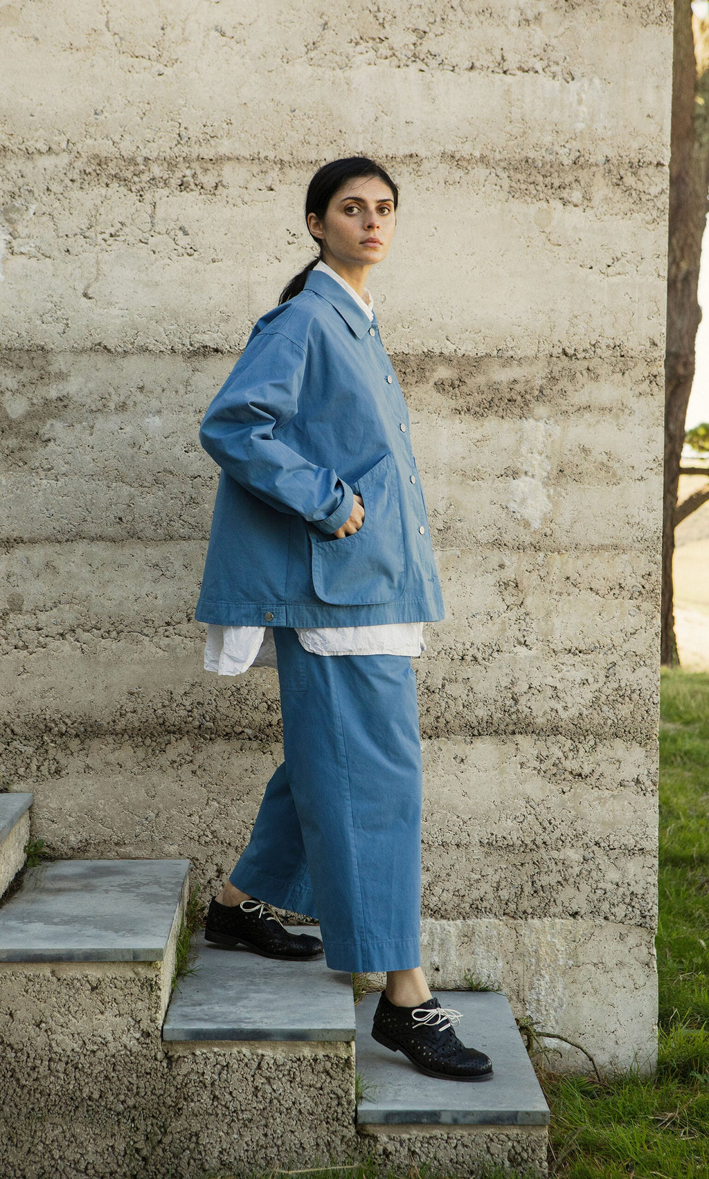 Plümo- Wearing the Flanders Jacket in French Blue, a person stands on outdoor steps by a textured concrete wall.