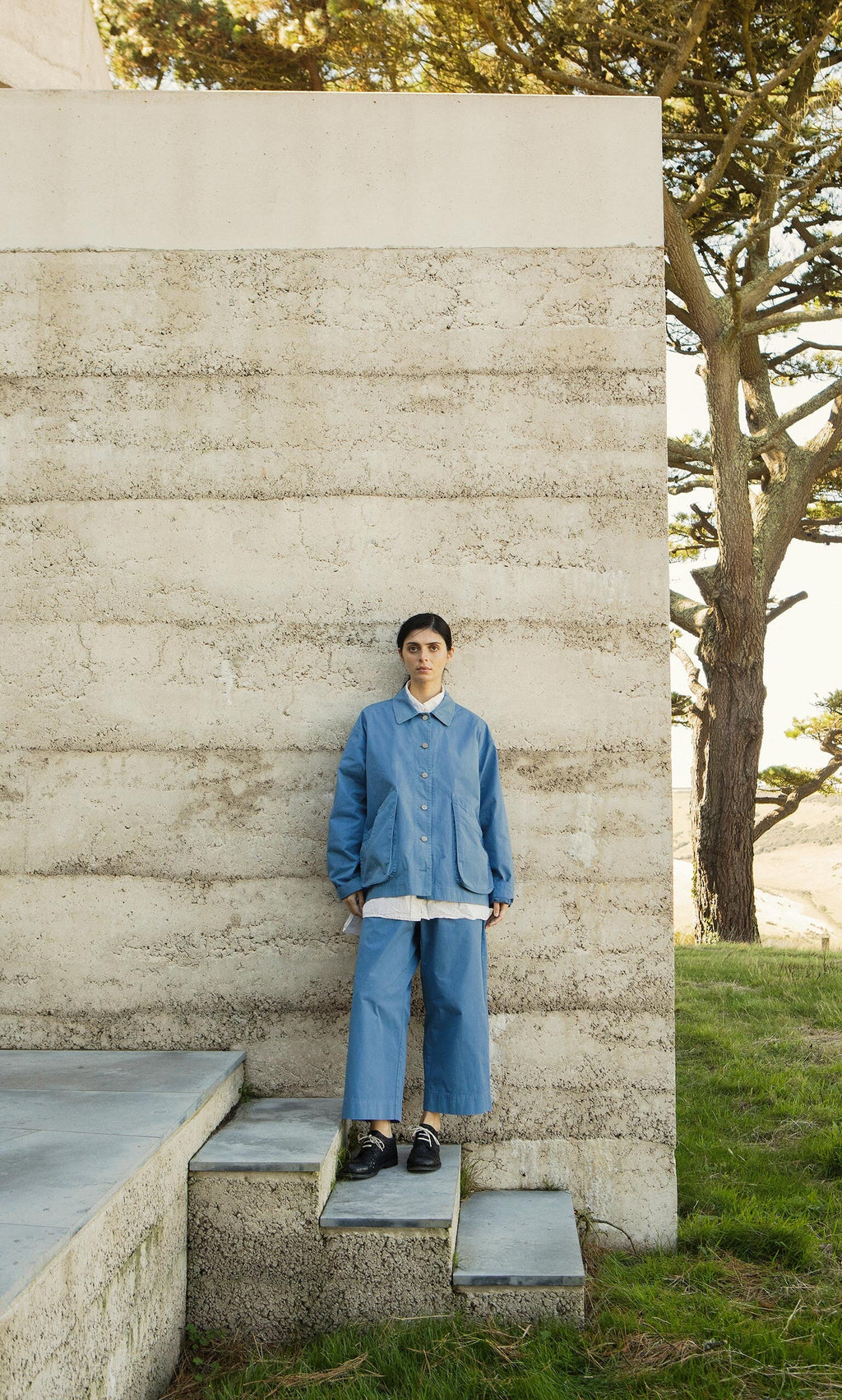 Plümo- Model wears Cromer Pants in French Blue, made in London, standing by a concrete wall with grass and trees behind.
