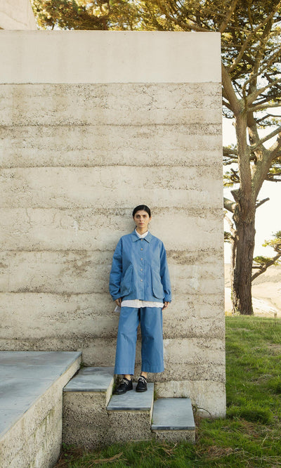Plümo- Model wears Cromer Pants in French Blue, made in London, standing by a concrete wall with grass and trees behind.