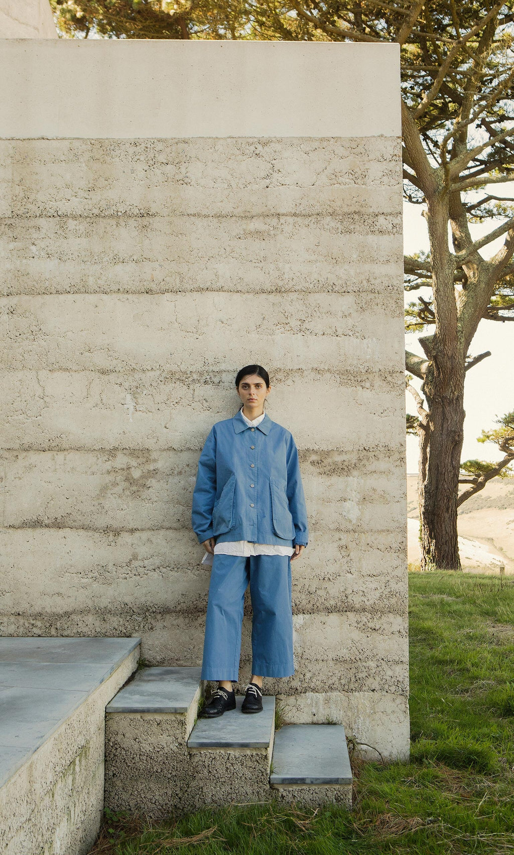 Plümo- Someone in a Flanders Jacket - French Blue stands on steps beside a textured concrete wall, grass, and a tree.