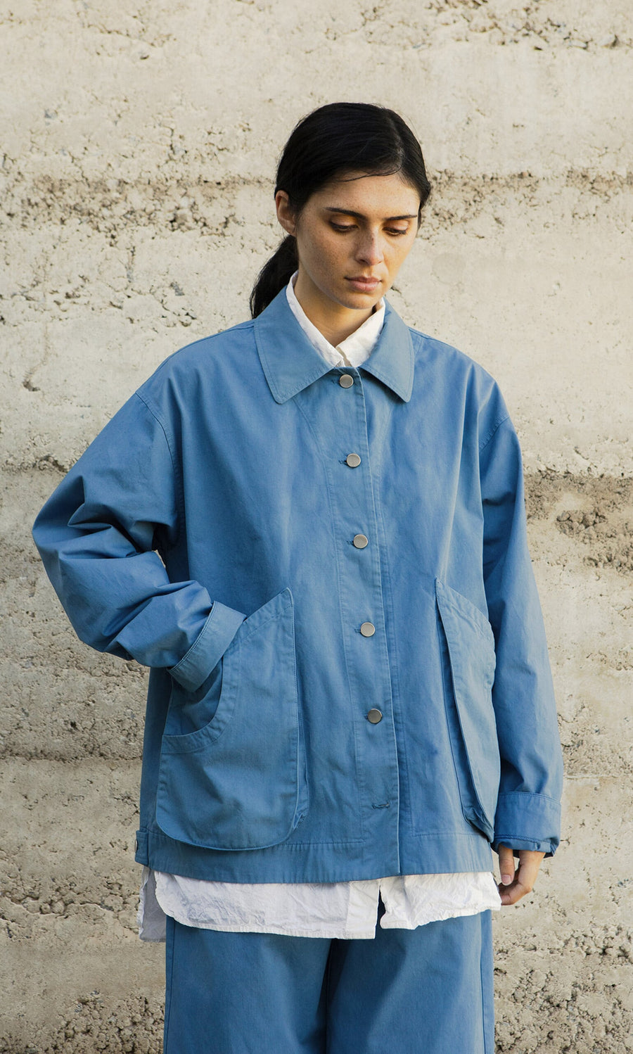 Plümo- A person in a Flanders Jacket - French Blue stands by a beige wall, looking down with a neutral expression.