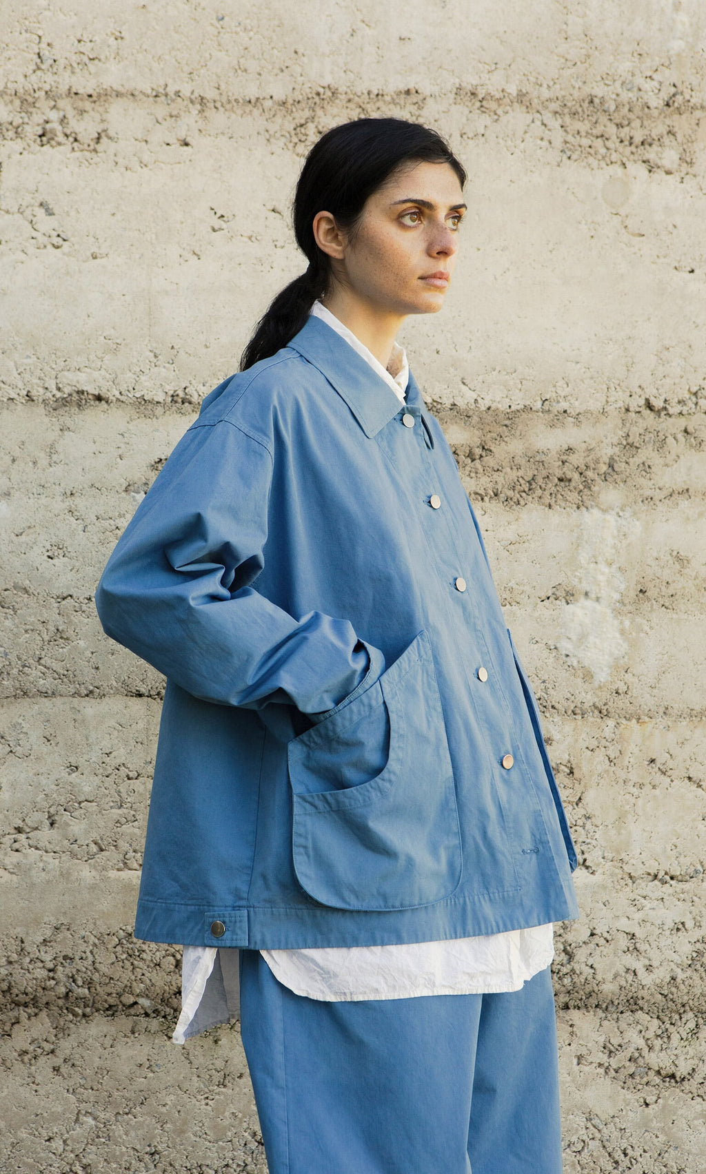 Plümo- Person wears the Flanders Jacket in French Blue over a white shirt, standing by a textured concrete wall.