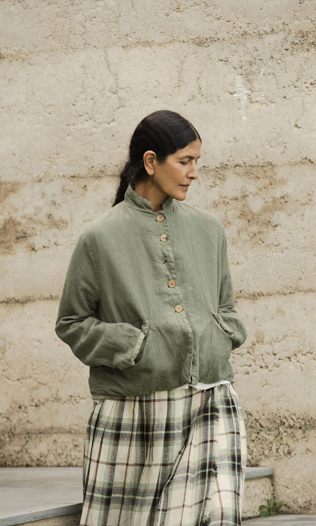 Plümo- Woman in the Frances Jacket stands outdoors, hands in pockets, looking down against a textured wall.
