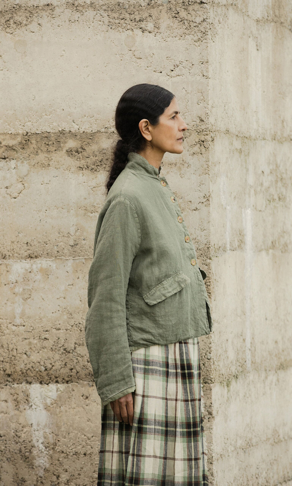 Plümo- Woman in a Frances Jacket and plaid skirt stands sideways by a beige concrete wall.