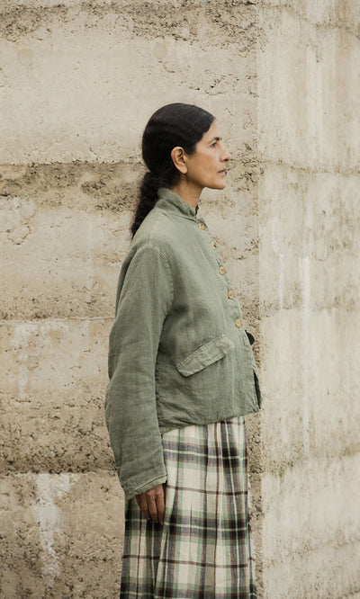 Plümo- Woman in a Frances Jacket and plaid skirt stands sideways by a beige concrete wall.
