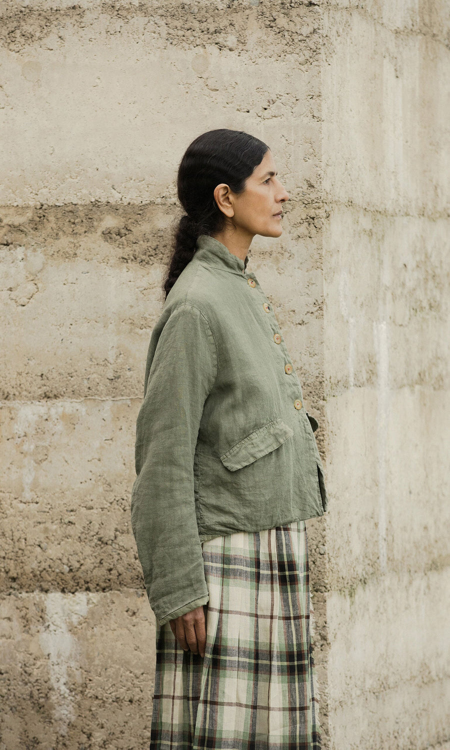 Plümo- Woman in a Frances Jacket and plaid skirt stands sideways by a beige concrete wall.