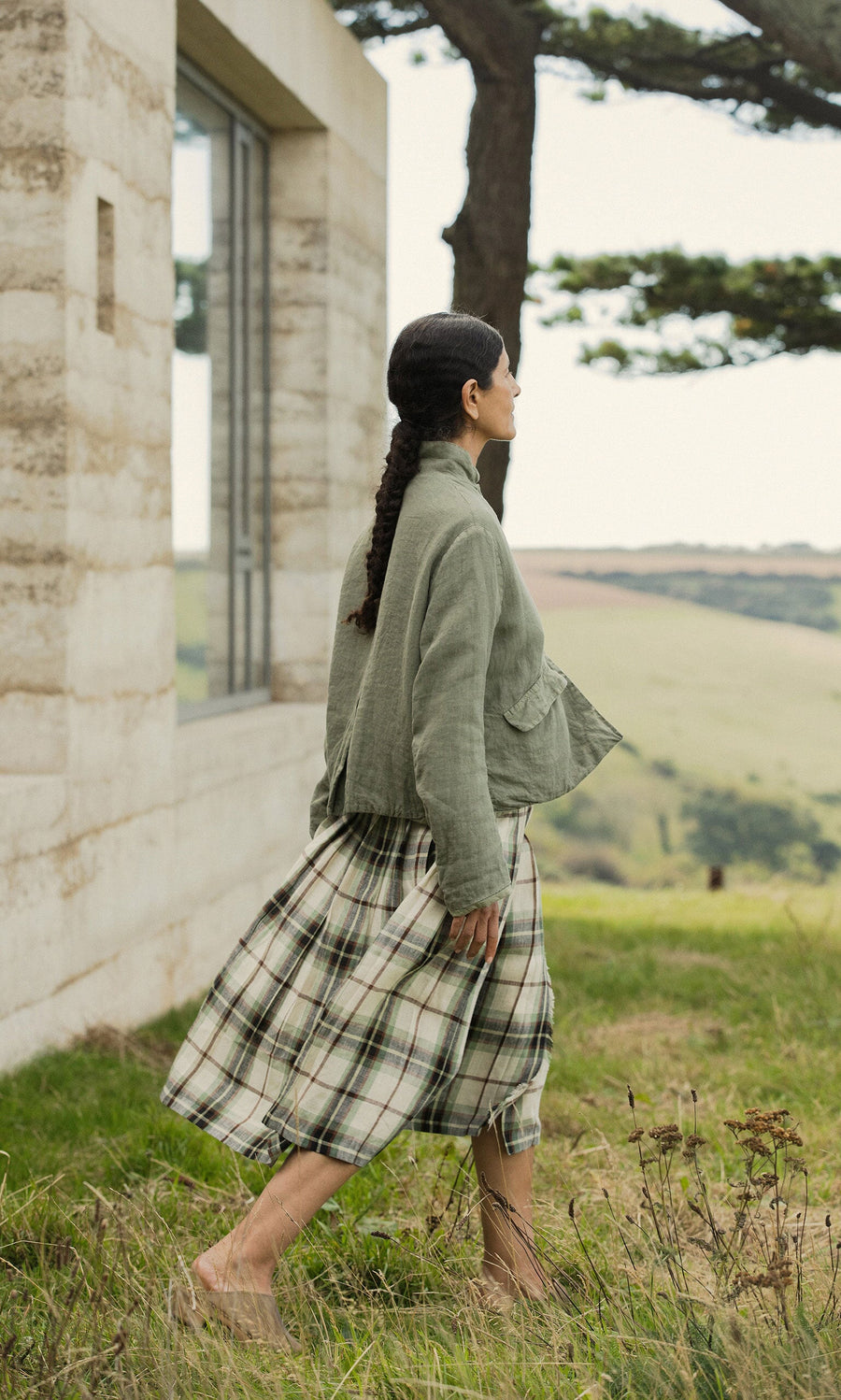 Plümo- A woman in a Frances Jacket and plaid skirt walks outside near a modern building with hills and trees behind her.