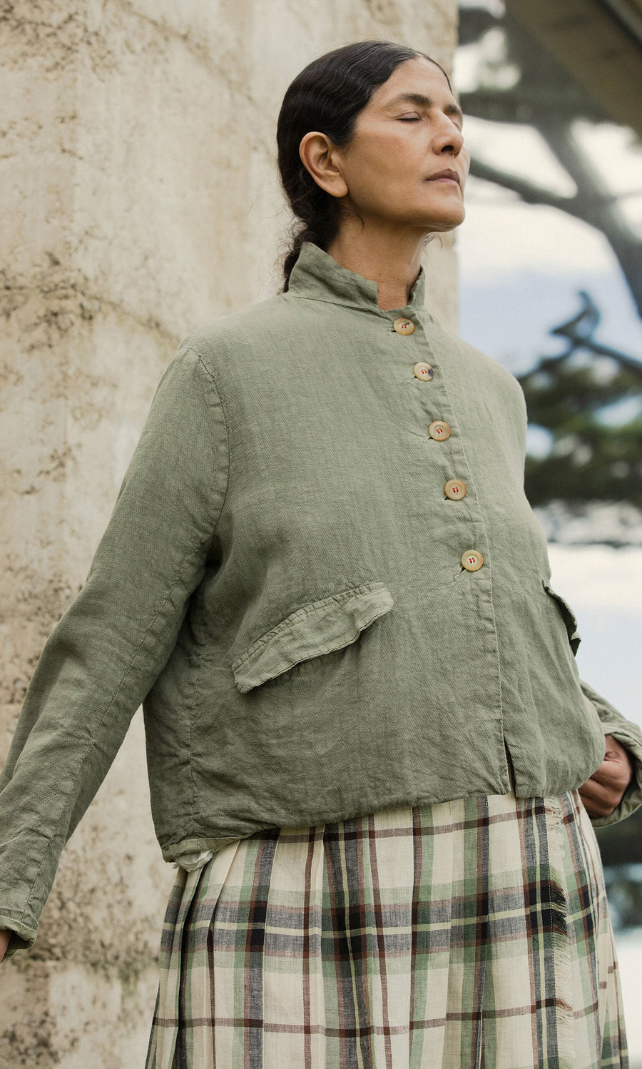 Plümo- A woman in a Frances Jacket and plaid skirt stands outdoors with her eyes closed, leaning on a stone wall.