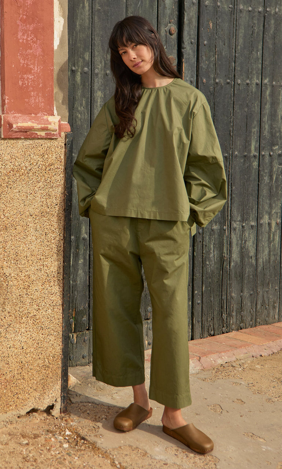 Plümo- Woman in the Gail Blouse - Calke Green and pants with brown slip-ons, standing by a rustic wooden door.