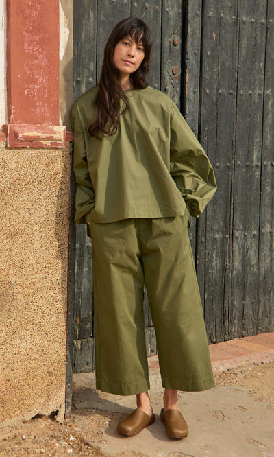 Plümo- Woman in the Gail Blouse - Calke Green and matching pants stands by a rustic wall, wearing brown slip-on shoes.
