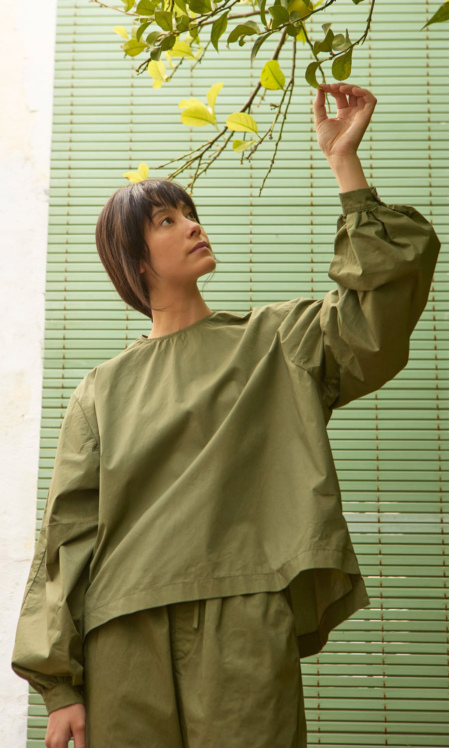 Plümo- Person in the Gail Blouse - Calke Green reaches up toward a leafy branch against a green slatted background.