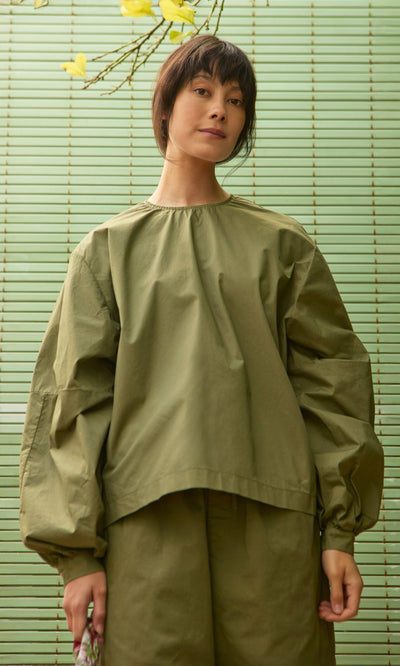 Plümo- Model wearing Gail Blouse in Calke Green stands before striped background with yellow flowers above.