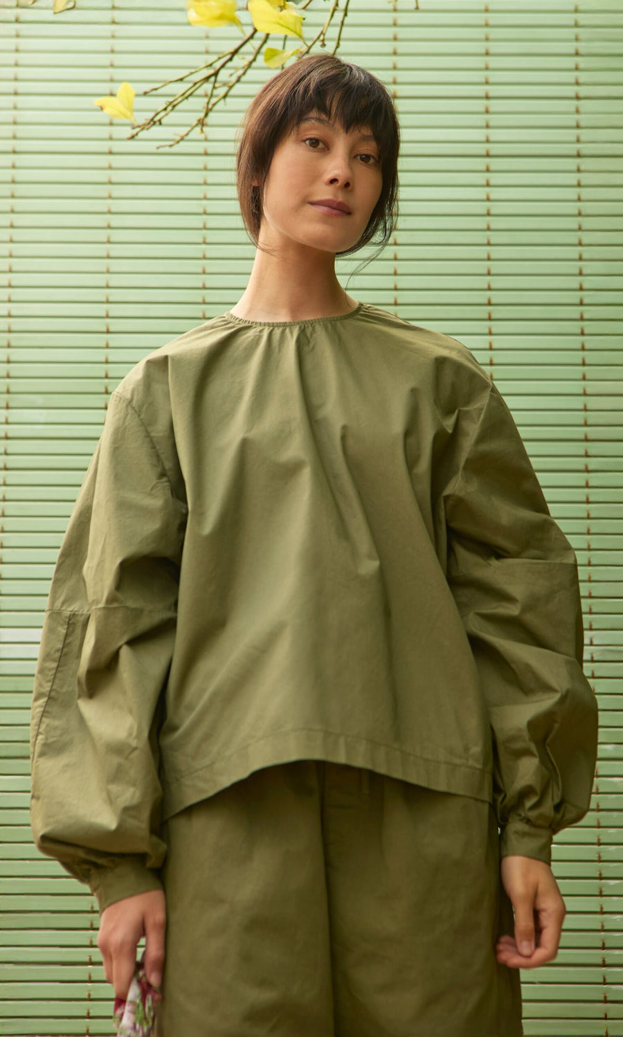 Plümo- Model wearing Gail Blouse in Calke Green stands before striped background with yellow flowers above.