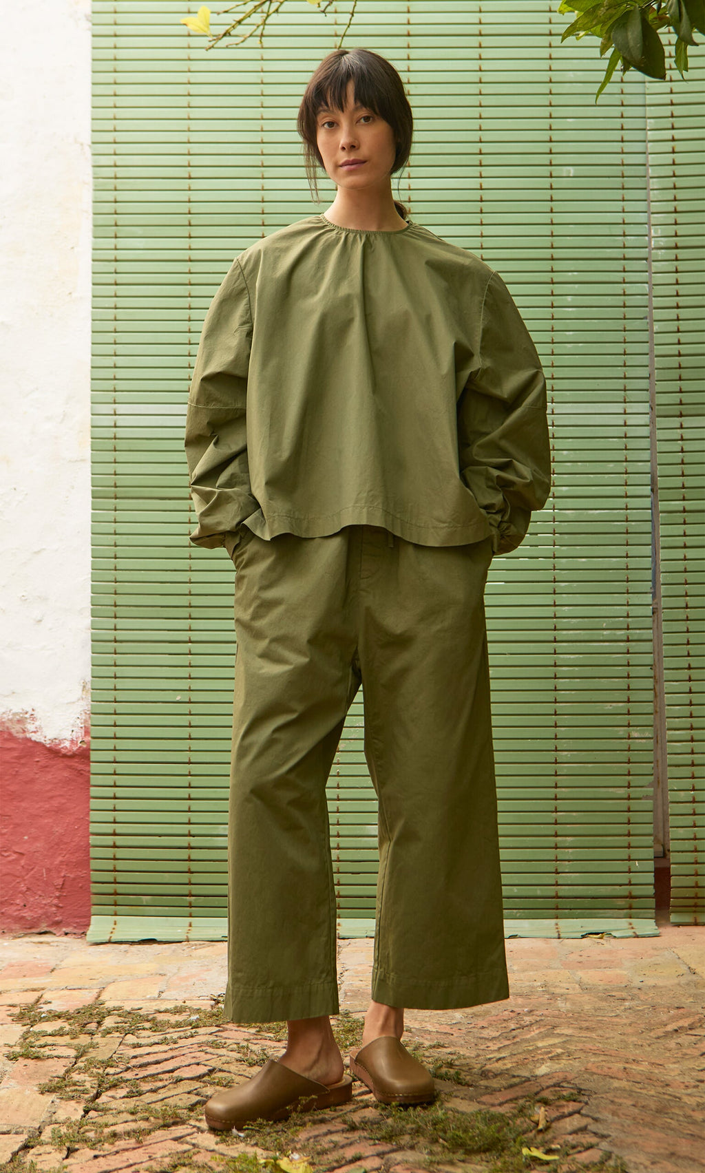 Plümo- Person in loose olive-green cotton poplin top and Calke Green Pants, standing by green blinds on a rustic patio.