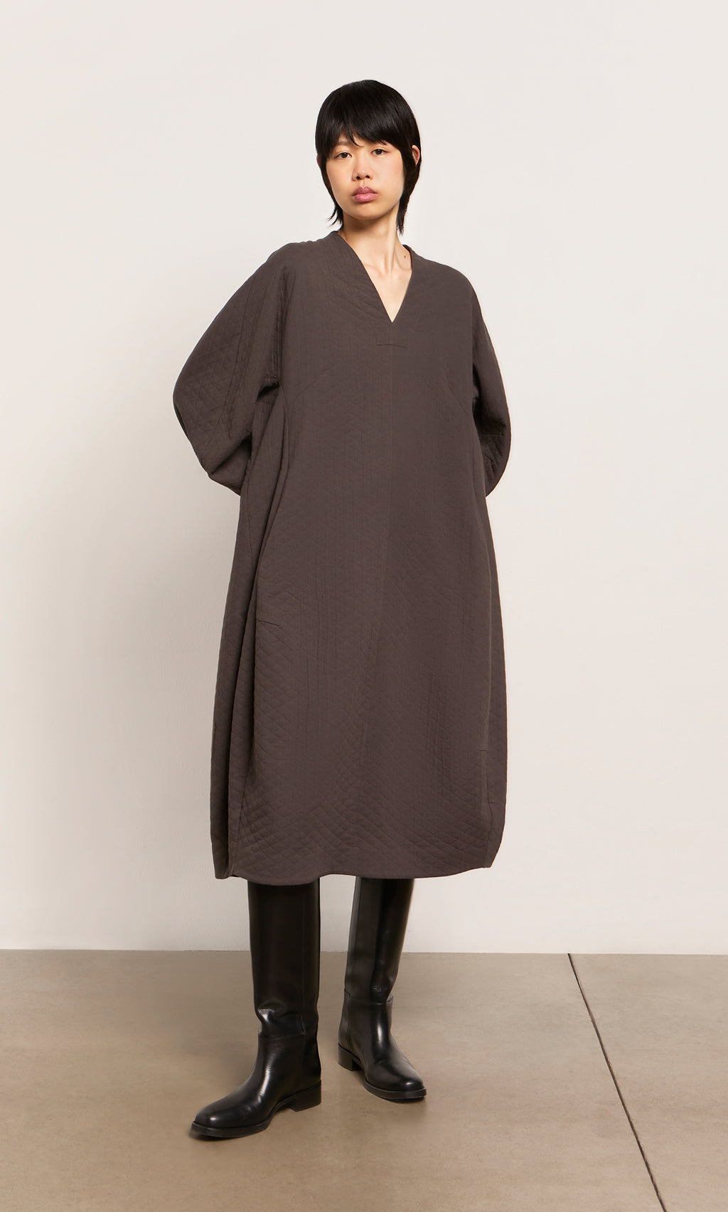 Plümo  Person wears a loose, dark brown Grenyer Dress from Plümo Studio and black knee-high boots against a plain background.