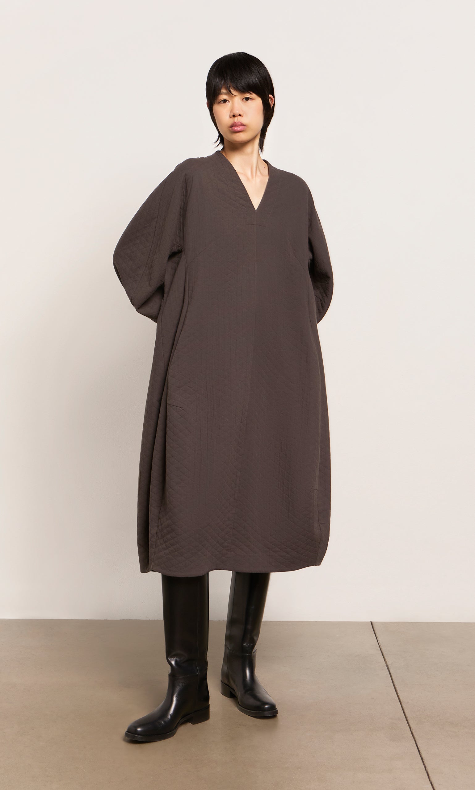 Plümo  Person wears a loose, dark brown Grenyer Dress from Plümo Studio and black knee-high boots against a plain background.